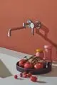 Wandmontierter Küchenarmatur in chrom mit einem schlichten Design, installiert an einer roten Wand. Dazu ein starker Kontrast mit einem weißen Tisch und frischen Lebensmitteln. Ideal Standard.